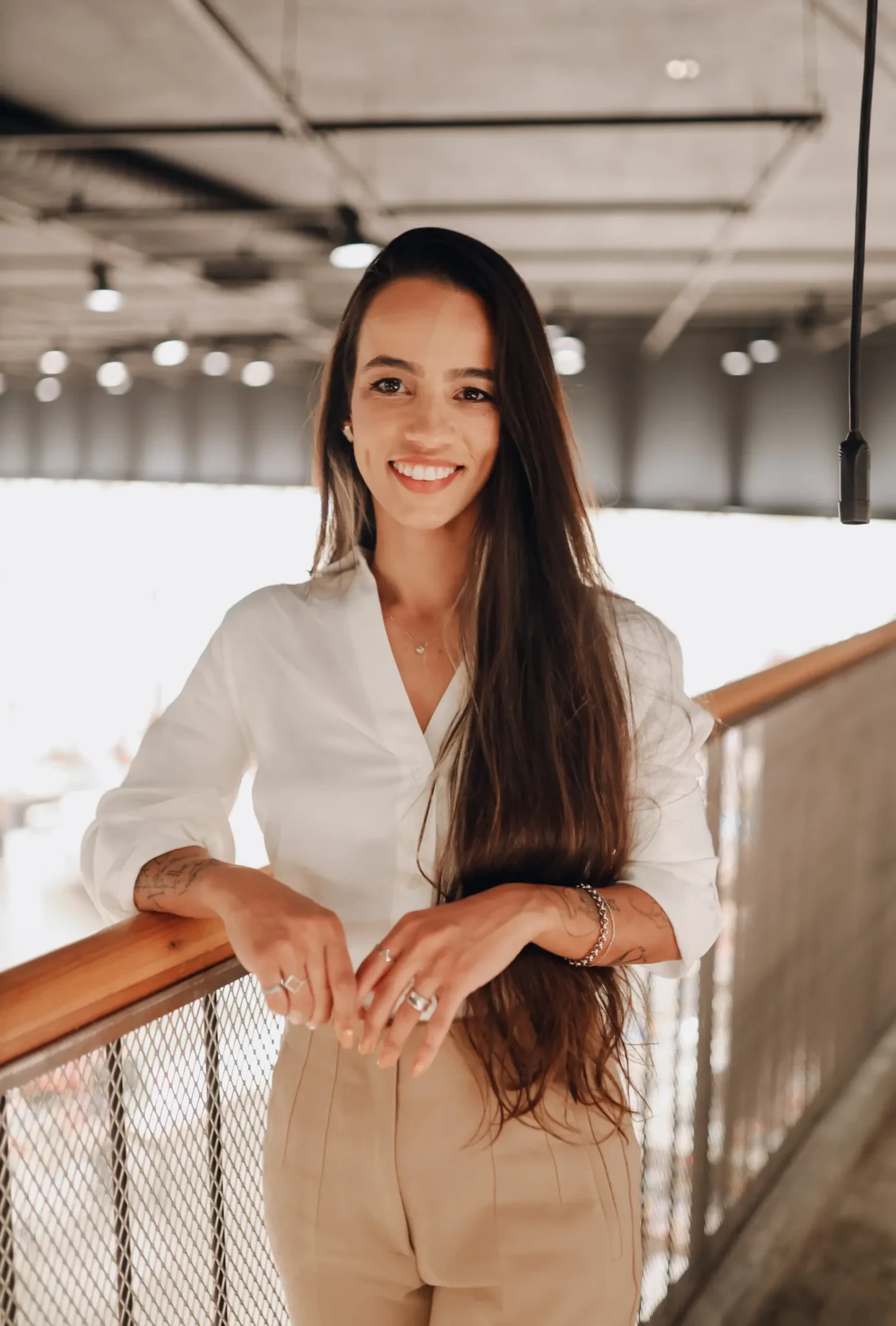 Monique está em pé em um ambiente industrial, apoiando seu braço no parapeito do andar superior. Monique está sorrindo e veste roupa social clara